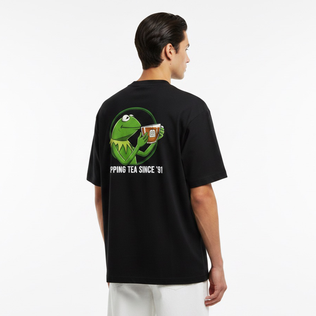 Person wearing a white t-shirt with a graphic of a green frog holding a cup and text on the back.